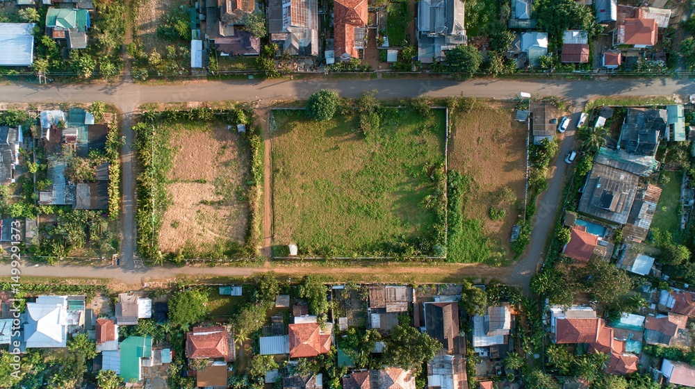 Obraz premium Aerial view of plots of land in a neighborhood setting, with houses and trees surrounding open grassy and dirt areas, divided by a road.