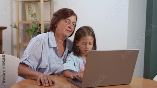 Little girl sitting with grandmother using laptop at home, both exploring digital world together while sharing a learning moment in a warm and engaging atmosphere