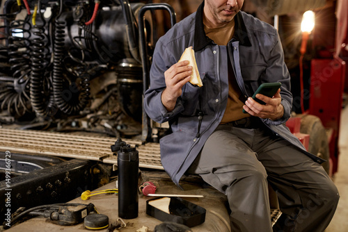 Mechanic sitting in workshop taking break by eating sandwich and using phone. Surrounded by various tools and equipment in industrial setting