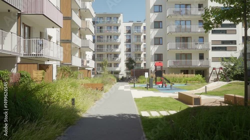 A modern gated residential complex featuring multi-family apartment buildings, a children's playground, landscaped recreational areas with benches, and pedestrian walkways in a sunny summer day.