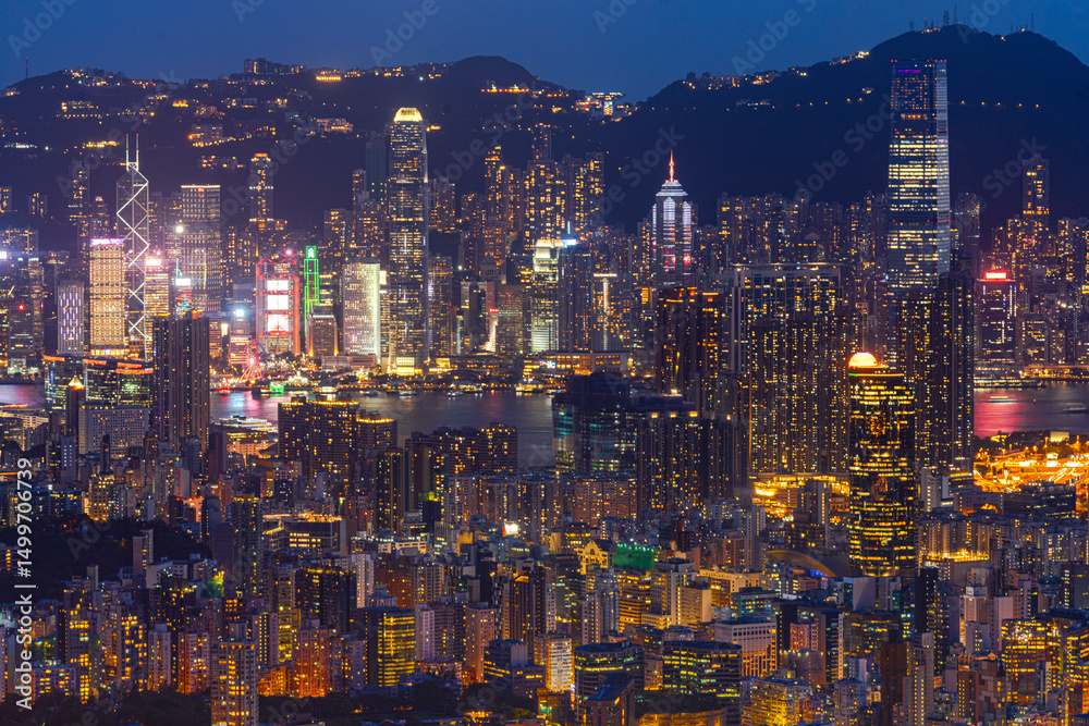Obraz premium Hong Kong city view from above with modern buildings and towns lit up at night across island. Light hits buildings which creating focal point in city with modern buildings and road with traffic light.