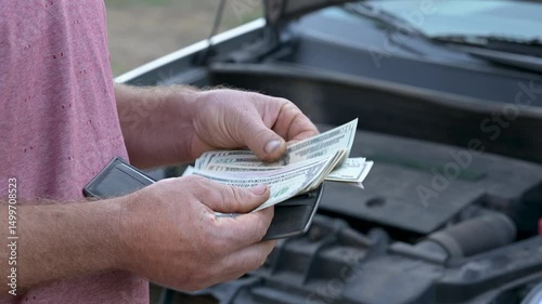 Counting dollars cash for car repair payment near the open hood. A person counts small banknotes near an open car hood, paying for repairs with his last money