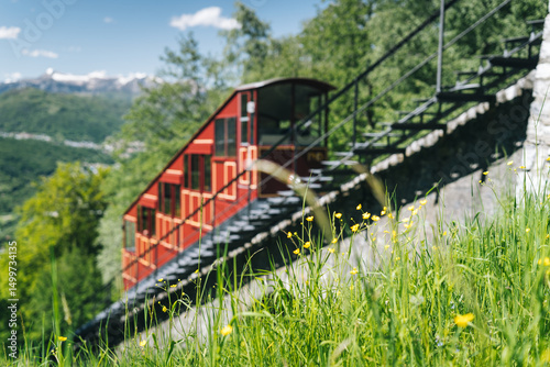 Funicular tram ascending steep rail tracks up mountain on sunny day, green forest and mountains in the distance