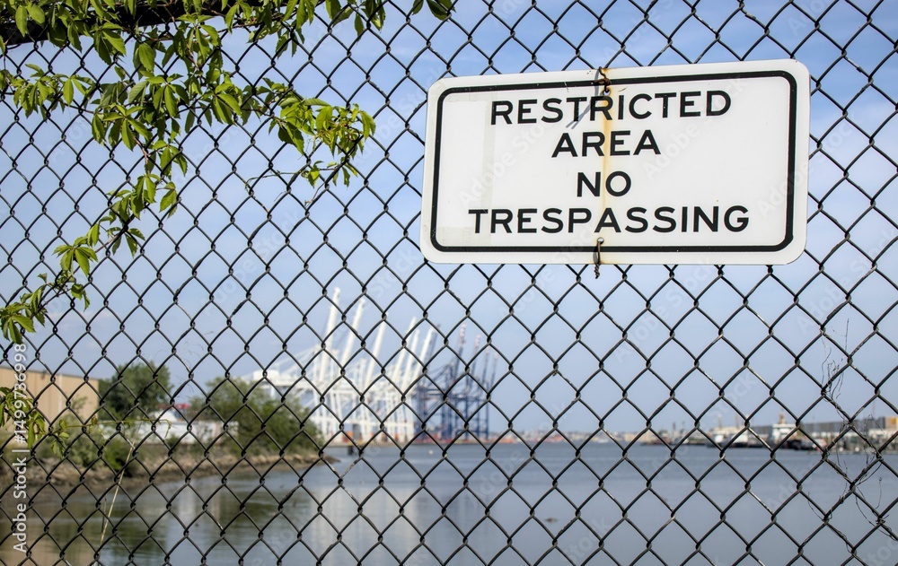 Naklejka premium restricted area no trespassing sign on black metal chain link fence in front of shipping docks in bayonne new jersey (infrastructure industrial ship container delivery security international tariff)