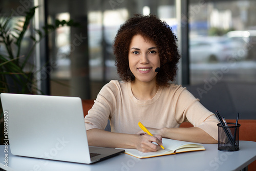 Black beautiful woman smiling during business phone call. African-American woman wearing headphones talks online with colleagues or clients using a computer application for video communication
