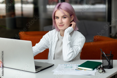 Young modern woman freelancer with pink hair working remotely using laptop and sitting in coworking space or home
