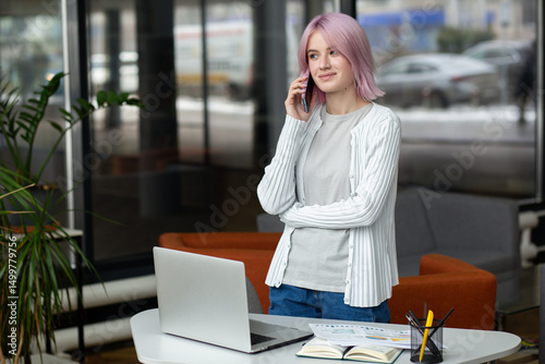 Happy young woman talking on the mobile phone and smiling while standing at her working place in office. Pink haired young modern inreterpreneur