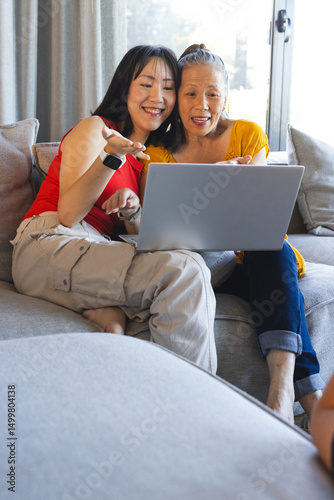 Smiling Chinese mother and adult daughter sitting on gray sofa in modern living room, using laptop