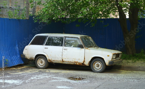 An old white rusty car is parked near a blue fence, Stakhanovtsev Street, Saint Petersburg, Russia, May 22, 2025
