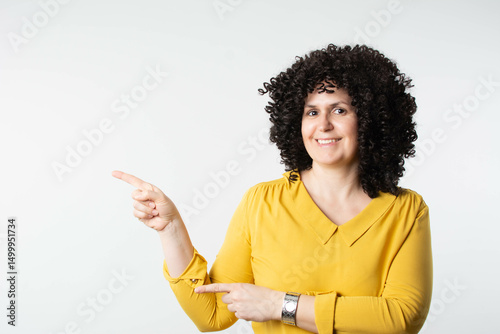 Beautiful mature woman with curly hair, smiling and pointing away. She stands against a bright background, radiating confidence and happiness. 