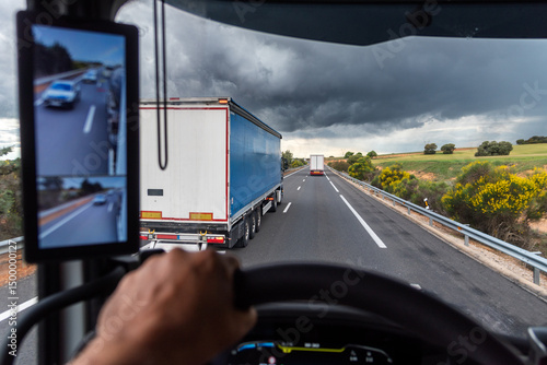 View from inside a truck cabin as another truck overtakes on the road, with a dark, storm threatening sky overhead.