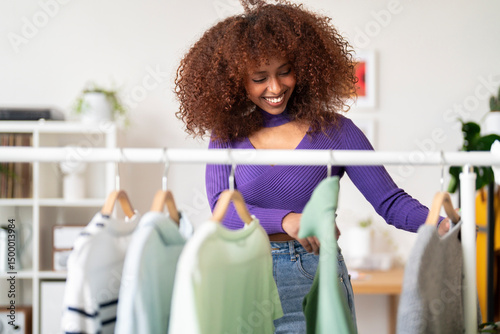 A smiling woman with curly hair is happily looking through clothes hanging on a rack. She is likely deciding what to wear for the day or organizing her wardrobe.