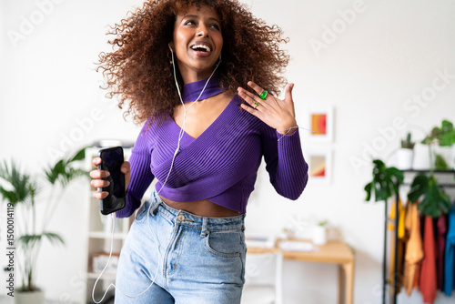 A joyful woman with curly hair is dancing and listening to music through wired headphones connected to her phone, expressing happiness and freedom in her home.