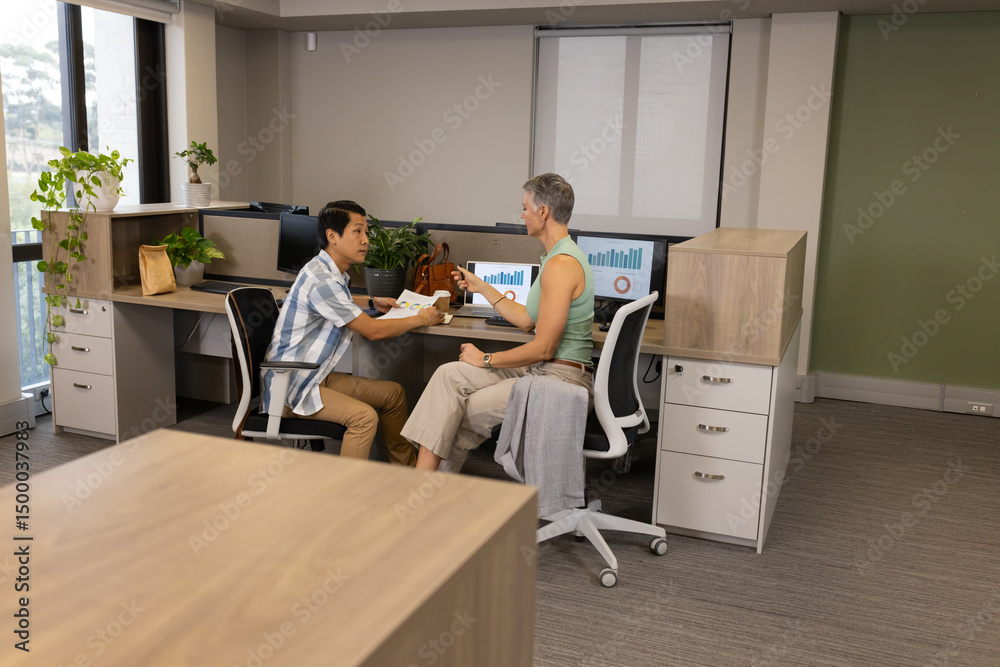 Diverse coworkers discussing reports at shared desk in modern office, with dual monitors Diverse coworkers discussing reports at shared desk in modern office, with dual monitors