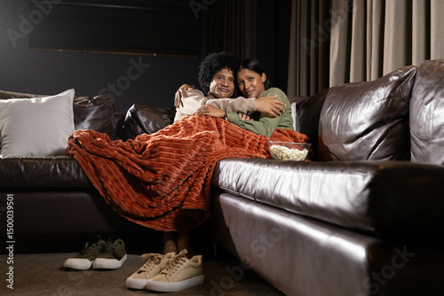 Cuddling diverse couple snuggling under rust-colored blanket on leather sofa at home, with popcorn