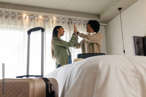 High-fiving Diverse friends standing beside bed in hotel room, with rolling suitcase and travel bag