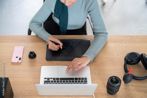 A woman in a hijab uses a drawing tablet and laptop for creative work. She is likely designing or editing, utilizing digital tools for her projects.