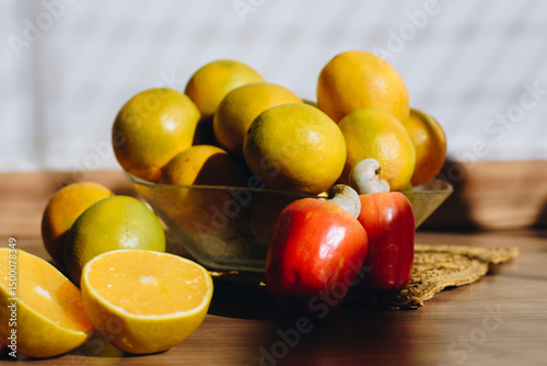 Fruit bowl with oranges on a wooden table under sunlight. Nearby, cashew fruits and a halved orange. Natural light casts soft shadows, highlighting the fresh fruits' colors and textures.