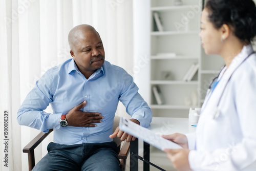 Black male patient holding stomach, suffering acute abdominal pain discomfort during meeting with female doctor in bright modern clinic, mature man feeling unwell, closeup