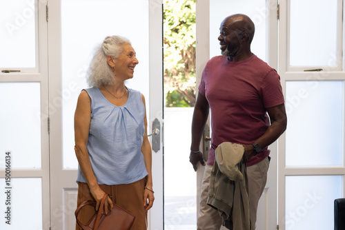 Standing senior couple chatting in home entryway, with brown leather handbag and light jacket photo