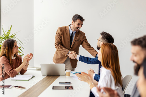 Chief is shaking hands with successful employee at boardroom while managers applauding them.