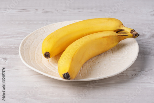 Yellow bananas on a white ceramic plate resting on a wooden table in a bright kitchen setting near a window