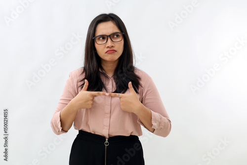 Young Asian woman standing with hesitant and uncertain gesture and expression keeps pointing her index finger, glancing sideways, thinking about something, wearing formal shirt, with white background.