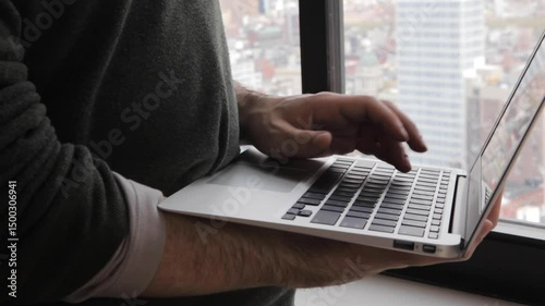 Handheld close-up of fingers typing on a laptop by a window overlooking Brooklyn and Manhattan below  and in the background. 