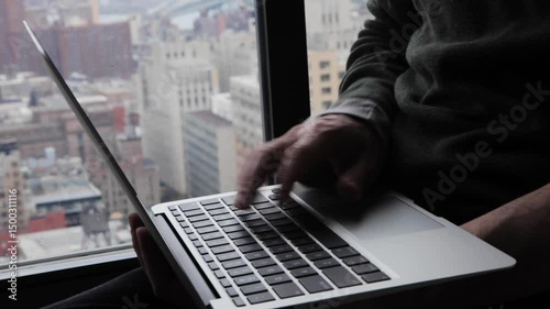 Handheld close-up of fingers typing on a laptop by a window overlooking Brooklyn and Manhattan below  and in the background. 