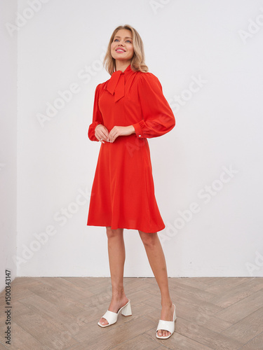 Smiling woman in red dress standing and holding hands together, wearing white heeled sandals. Blonde hair, long sleeves, tie collar. Light wooden floor with modern, minimal style