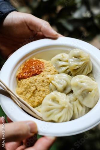 A plate of steamed chicken momos held in a man’s hands, served with spicy chutney, wooden fork and knife at a Nepalese street food stall in Little India - Harris Park, Sydney, Australia 