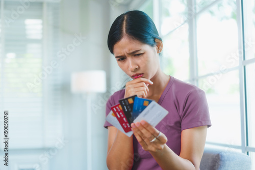 Stressed Asian woman sits on a sofa holding several credit cards, feeling anxious and overwhelmed by debt and unpaid bills, reflecting financial pressure, poor budgeting,  emotional distress at home