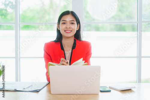 Confident Asian businesswoman sits at a modern desk  reviewing financial charts and planning reports. Surrounded by a laptop and plant  she showcases focus  organization  and professionalism