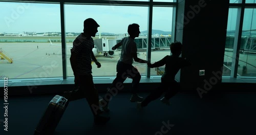 Silhouetted shot of family energetically running through airport terminal, with young boy leading and father with suitcase trailing behind. Passengers hurrying toward boarding gate in funny rush
