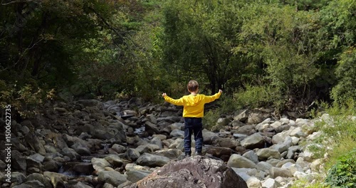 Jubilant boy in yellow hoodie stands triumphantly on large rock in mountain riverbed, shaking his arms with excitement after overcoming obstacles.