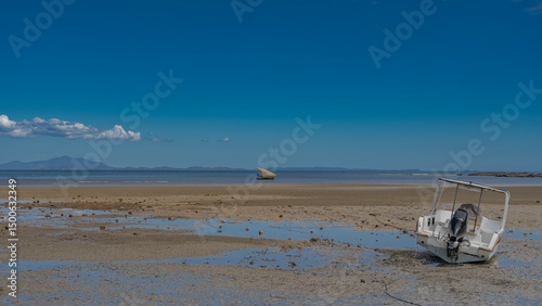Low tide in the ocean. A motorboat tilted on the exposed sandy seabed. Puddles of water. There's a sailboat in the ocean. Mountains on the horizon. Clear blue sky. Madagascar. Nosy Be. Copy space.