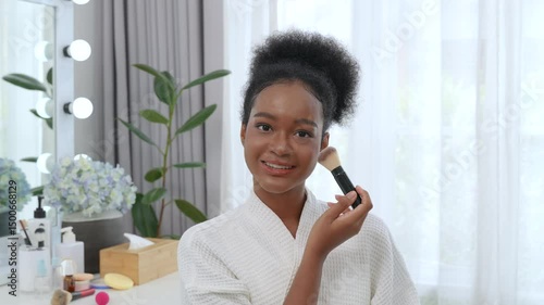 Beautiful black teenage girl using blusher to apply makeup in front of a mirror at home.