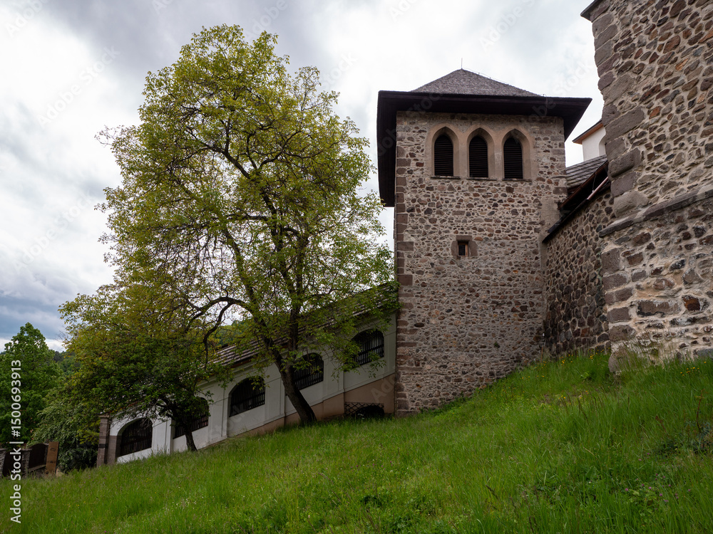 Fototapeta premium Covered staircase leads to the fortified entrance of Kremnica Castle, passing under trees and beside medieval stone walls and a watchtower, surrounded by lush greenery.