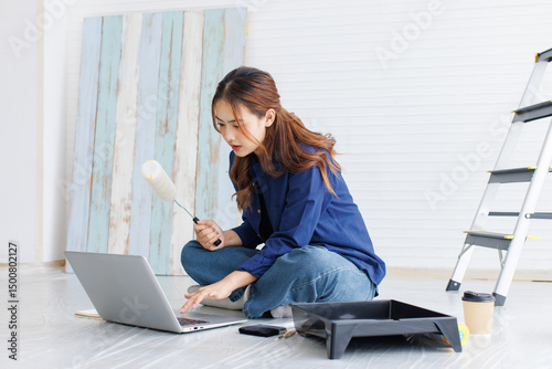 Asian young beautiful woman long curly hair wearing blue shirt jean sit on floor look at laptop computer learning before repairing room painting wall house or apartment rollers brush ladder background