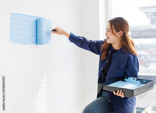 Asian young beautiful woman smiling cheerful long curly hair wearing blue shirt jeans sitting on ladder painting wall with roller brush light blue for new home repair renovation decoration concept.