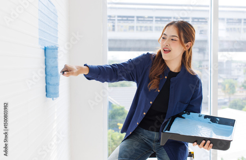Asian young beautiful woman smiling cheerful long curly hair wearing blue shirt jeans sitting on ladder painting wall with roller brush light blue for new home repair renovation decoration concept.