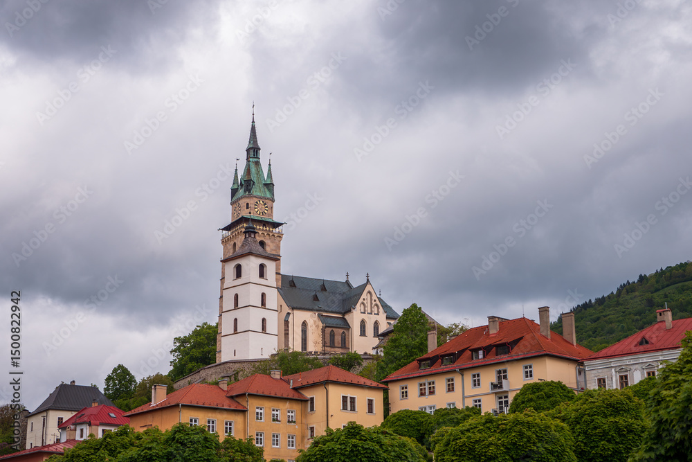 Fototapeta premium Dominant view of Kremnica Castle with the Church of St. Catherine and its iconic clock tower, rising above colorful townhouses and nestled in the green Štiavnica Hills.