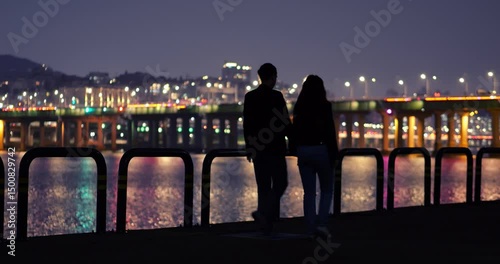 Silhouetted tourist couple walking hand in hand along Han River at late hour, blurred city lights and glowing Hannam Bridge in background. Nighttime stroll as young man and woman share quiet moments