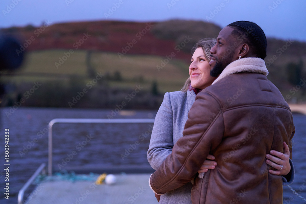 Fototapeta premium Smiling couple in warm clothing embracing on pier at dusk