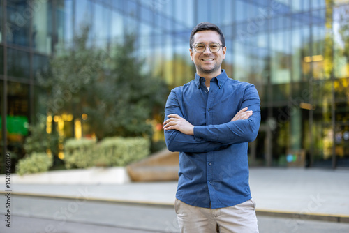 Obraz na plátně A confident businessman, smiling with arms crossed, stands confidently in front of a modern office building on a sunny day
