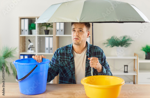 Man troubled at home holding umbrella among plastic bucket and basin, hole, crack in ceiling, water flowing out of roof, need of professional roofer, sad with necessary room repairs to find, fix leaks