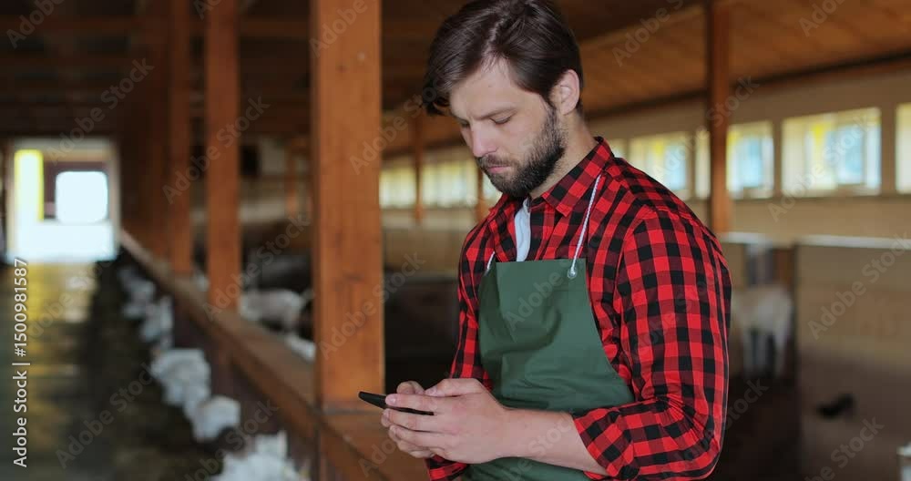 Portrait of young male farmer sitting in goat stable and texting message on smartphone. Man farmer tapping and scrolling on mobile phone. Modern livestock farming business.