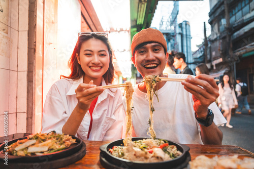 Asian couple man and woman enjoy eating street food noodle at night market. Traveler Asian couple blogger Happy tourists with Traditional food local road at thailand bangkok city.