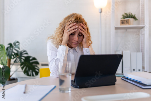 Stressed businesswoman holding head in hands while working on laptop at desk in modern office, facing work pressure or burnout.