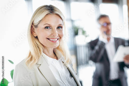 Smiling businesswoman posing in a modern office environment with a businessman coworker in the background making a call Free Download Smiling businesswoman posing in a modern office environment with a businessman coworker in the background making a call - GFXLoad